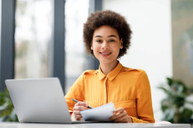 Çevrimiçi Webinar öğrenen, not yazan, üniversite kütüphanesinde okuyan, masada oturan ve kameraya gülümseyen pozitif siyah öğrenci kız.