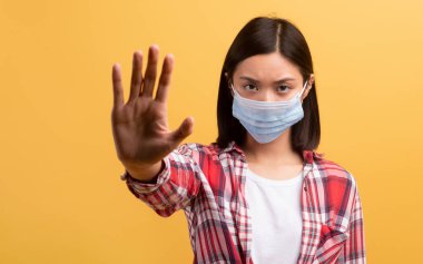 Koruyucu maskeli, elinde dur işareti olan, sarı arka planda izole edilmiş, stüdyo fotoğrafı olan kendine güvenen Japon bir kız öğrenci. Sağlık sorunları, hastalıklar, virüsler