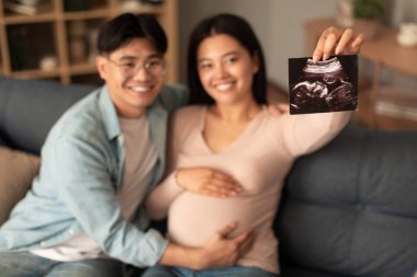 Neşeli hamile Koreli genç eşler ultrason embriyo fotoğrafı gösteriyorlar, bebek bekliyorlar ve evde kanepede oturmuş kameraya gülümsüyorlar. Sonogram resmine seçici odaklanma