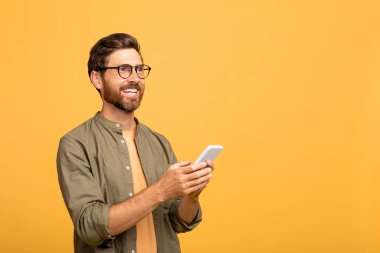 Chatbot, online asistan konsepti. Neşeli Avrupalı orta yaşlı adam cep telefonu kullanıyor ve fotokopi alanına bakıyor, sarı stüdyo arka planında izole edilmiş.