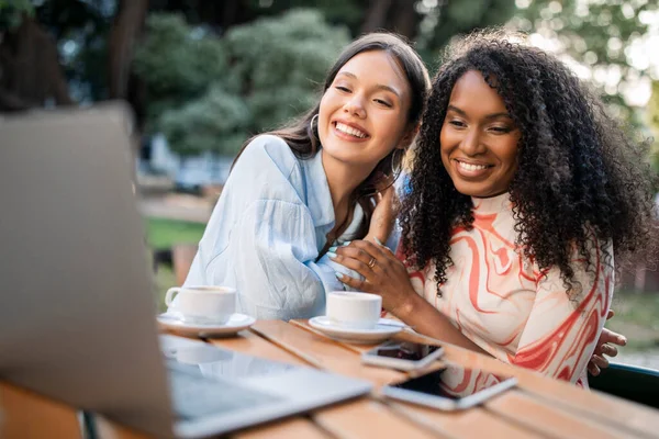 Portrait of happy young multiethnic female friends using laptop in cafe outdoors, cheerful beautiful besties watching videos on computer or shopping online, enjoying spending time together outside