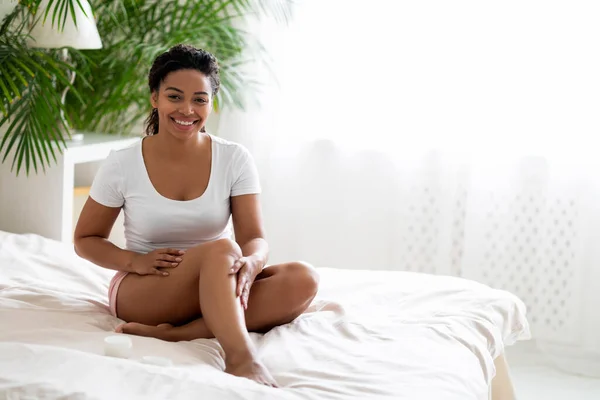 Happy young black lady applying moisturizer cream on legs while sitting on bed at home, smiling african american female moisturizing skin after body shaving, making aftershave skincare routine