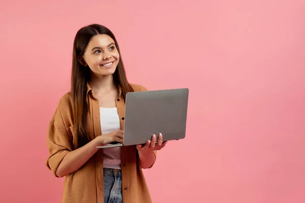 ラップトップを保持し コピースペースを脇に見て美しい笑顔のティーンガール オンライン教育や距離学習のためのコンピュータを使用して若い女性学生 ピンクの背景に立って コピースペース — ストック写真