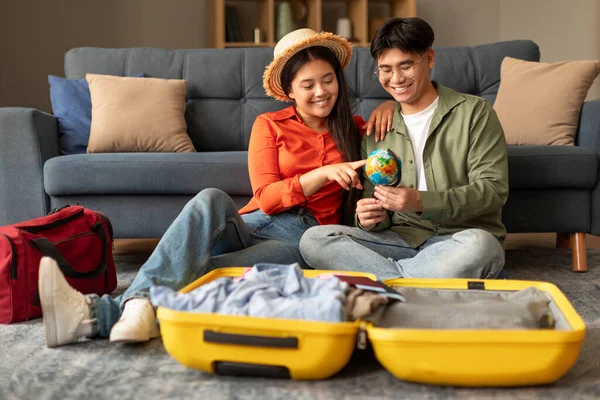 Vacation plans. Happy chinese couple planning travel, pointing at world globe choosing destination for holiday journey, sitting near suitcase preparing for trip at modern home interior
