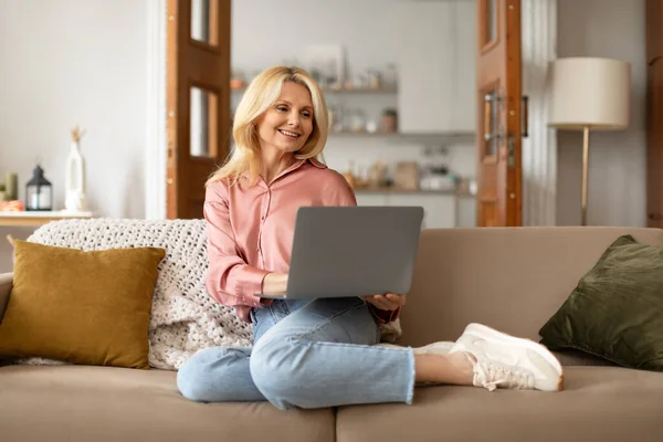 一位漂亮的老太太 穿着休闲装 坐在沙发上 坐在笔记本电脑上 在家里休息 上网看视频 在舒适的客厅里上网看博客 — 图库照片