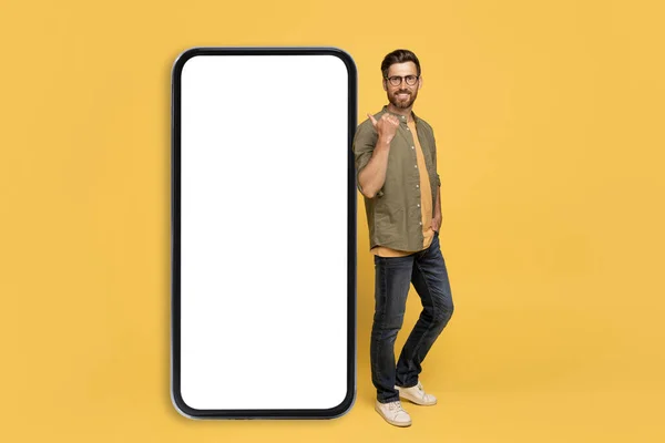 白い空白のスクリーンが付いている大きいスマートフォンを傾け 指さす陽気な中年の男 カメラで微笑み 広告のための自由なスペースを示し 黄色い背景の上に立って モックアップ — ストック写真