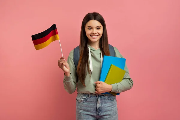 International Education. Beautiful Female Student With Backpack Holding German Flag, Portrait Of Smiling Teen Girl With Workbooks Posing On Pink Studio Background, Enjoying Study Abroad, Copy Space