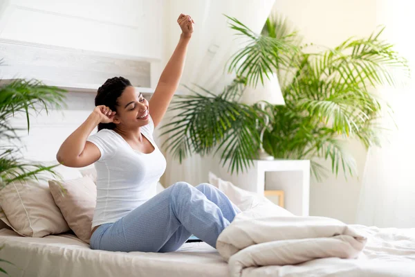 Beautiful Young Black Woman Stretching Hands While Sitting In Comfortable Bed, Portrait Of Happy African American Female Waking Up In The Morning, Relaxing In Cozy Bedroom, Enjoying Start Of The Day
