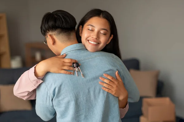Real Estate Dream Comes True. Happy Asian Couple Hugging, Wife Holding New House Keys, Celebrating Property Purchase At Home Interior, Standing Among Cardboard Moving Boxes