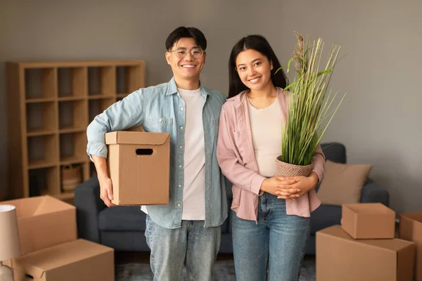 Family Moving House. Joyful Chinese Young Couple Posing With Packed Carton Boxes And Plant In A Pot, Ready For Relocation And Moving To New Home, Smiling To Camera. Own Apartment Property Purchase