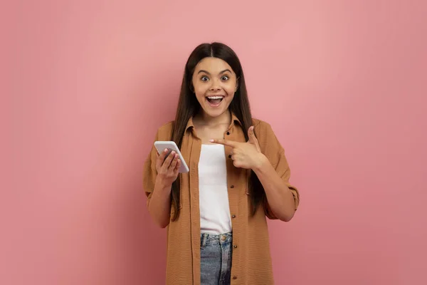 令人惊奇的报价 兴奋的少女手持智能手机指点 快乐的少女使用手机 在线成功 站在粉色背景下的工作室 复制空间 的刻画 — 图库照片