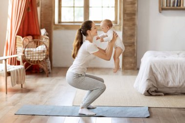 Bebekle spor aktiviteleri. Genç anne, küçük oğluyla evde egzersiz yapıyor. Kafkasyalı mutlu bir kadın kucağında bebekle yatak odasındaki spor paspası üzerinde çömelme antrenmanı yapıyor, boşluğu kopyalıyor.