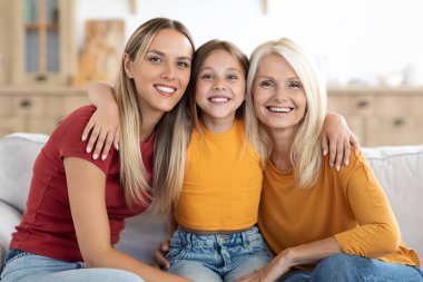 Üç nesildir mutlu kadın anne, kız ve büyükanne evdeki kanepede oturuyor, kameraya bakıyor, oturma odası arka planında sarılıyor ve gülümsüyor, kopya alanı, aile portresi
