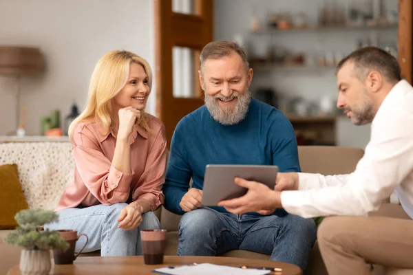 Agent Man Showing Digital Tablet To Excited Mature Clients Couple Offering His Digital Services, Sitting On Sofa In Their Living Room Indoor. Real Estate Buyers Choosing House Offers With Dealer