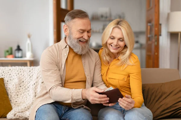 Financial Profit. Joyful Rich Senior Couple Holding Wallet Full Of Money Cash, Celebrating Great Income Sitting On Sofa In Modern Living Room At Home. Wealth And Family Budget Concept