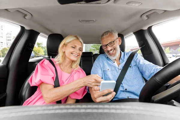 Happy senior man and woman having car trip together, using cell phone and smiling, copy space. Cheerful elderly couple travellers enjoying journey by cozy auto, tracking their way on smartphone