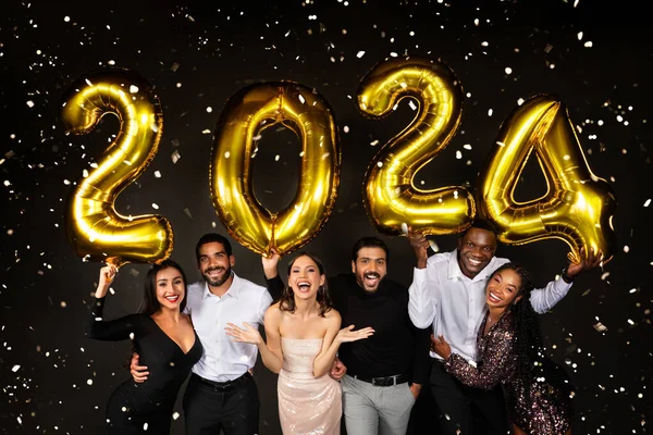 Happy multiracial men and women three loving millennial couples wearing nice outfits celebrating new year 2024 together on black background, holding golden number baloons and smiling at camera