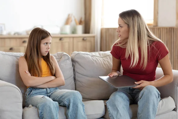 Strict mother scolding preteen daughter for long laptop use, angry woman with school aged girl sitting on couch in living room, holding computer in hands, parent reprimanding, lecturing child