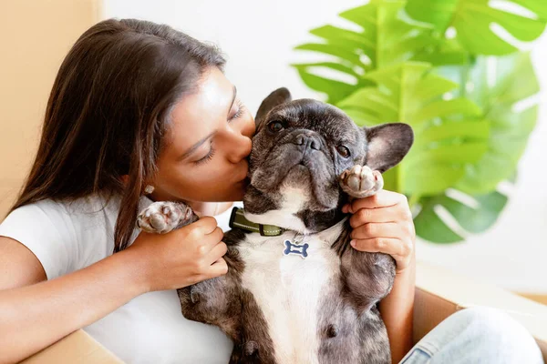 Closeup of pretty young woman with french bulldog in empty house among cardboxes with belongings, caressing kissing her pet dog in new apartment, enjoying new house. Rent, relocation, moving