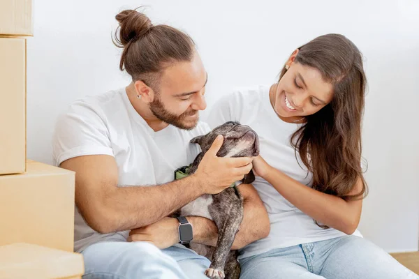 Home relocation concept. Positive young diverse couple stroking their dog in new home on moving day. Millennial multiracial family playing and cuddling with cute pet indoors, enjoying new apartment