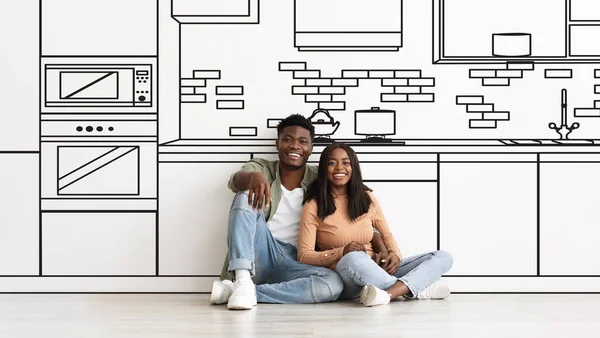 Real Estate, Family Housing. Happy Black Couple Hugging Near White Wall With Drawn Kitchen Interior, WIth Illustrations Of Modern Furniture, Sitting On Floor And Dreaming About Own House. Collage