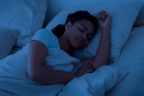 Healthy Sleeping Concept. Portrait of young black woman lying in bed with closed eyes in the night, calm african american female resting in bedroom, relaxing in dark room, smiling while having nap