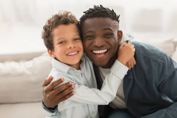 Happy Loving Family. African American Daddy Hugging His Little Son And Looking At Camera Indoors. Portrait Of Joyful Diverse Dad And Son. Fatherhood Joy, Child And Father Bond