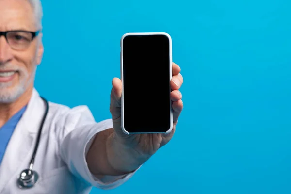Phone with black empty screen in smiling elderly doctor hand, mockup, copy space, blue studio background. Stay connected with your doctor. Telemedicine, e-health, online appointment concept