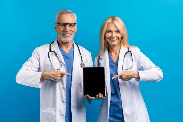 Online appointment with doc. Friendly senior doctors man and woman in medical white coats showing digital tablet with black empty screen, great mobile app for telemedicine, blue background, mockup