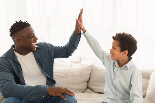 African American Dad and His Kid Boy Giving High Five, Celebrating Their Common Success, Having Fun And Spending Good Time Sitting On Couch At Home. Father And Son Bond, Togetherness Concept