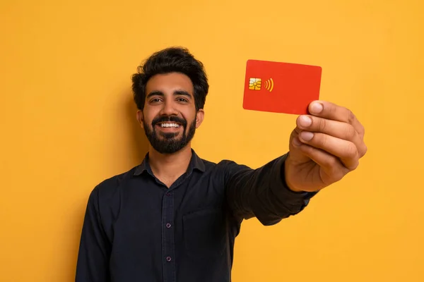 Cheerful Indian Man Showing His Credit Card To Camera, Advertising Great Bank Offer While Posing On Yellow Studio Background, Happy Guy Enjoying Easy Payments And Financial Safety, Closeup