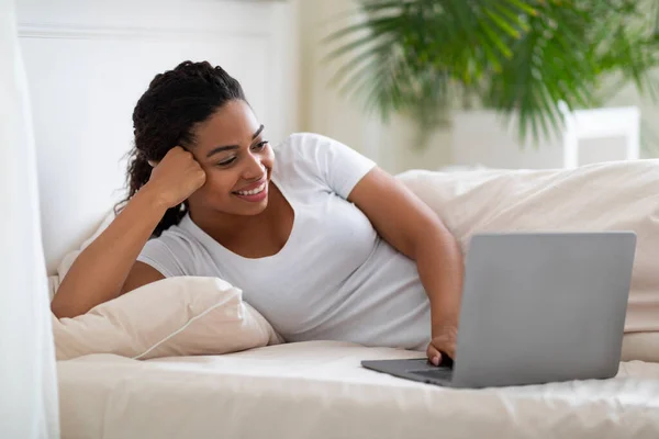 Portrait Of Young Black Female Relaxing With Laptop In Bed, Smiling Millennial African American Female Using Computer At Home While Relaxing In Bedroom, Watching Movies Or Browsing Internet