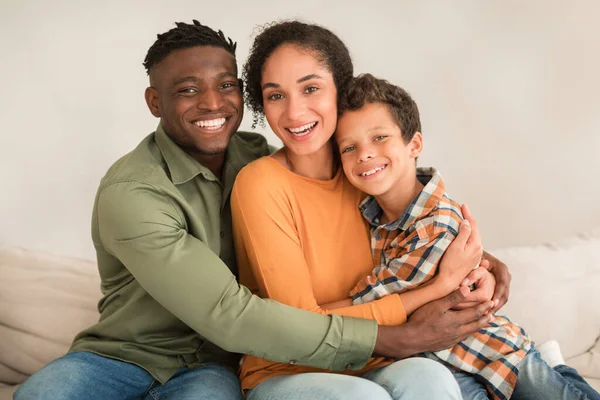 Diverse Family. Happy African American Man Hugging His Middle Eastern Wife And Little Kid Son Boy, Smiling To Camera Expressing Happiness Together, Sitting On Sofa At Home Interior