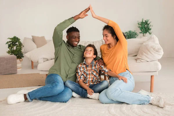 Real Estate Dreams. Multiethnic Father And Mother Joining Arms Like Roof Above Kid Son In Modern Living Room At Home, Sitting On Floor And Dreaming About New House Purchase. Family Housing Offer