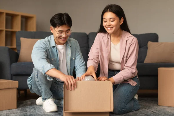 Moving New Home. Happy Korean Young Spouses Packing Carton Box, Preparing For Relocation At Modern Living Room Indoors. Asian Husband And Wife Putting Belongings In Boxes Renting New House
