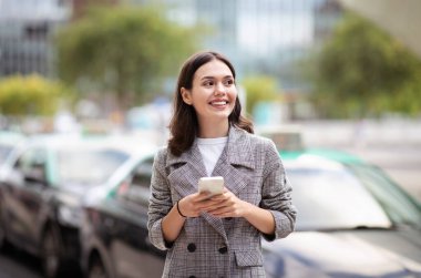 Mobil uygulama. Şehir merkezinde cep telefonuyla mesajlaşan genç bir iş kadını, akıllı telefondan taksi çağırıp cihazla iletişim kuruyor. Şehir hayatı, teknoloji.