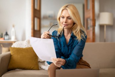 Dizüstü bilgisayarın yanında kağıt okuyan, elinde gözlük tutan, İç İşleri Bakanlığı 'nda modern bilgisayarlarda evrak işi yapan, iç mekanda koltukta oturan olgun bir kadın. İş Belgeleri, Muhasebe