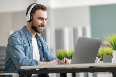 Çevrimiçi Eğitim. Kablosuz kulaklıklar takan ve not alan gülümseyen genç adam, Milenyum erkeği Webinar 'ı izliyor, İnternet dersine katılıyor, ofisteki masasında oturuyor.