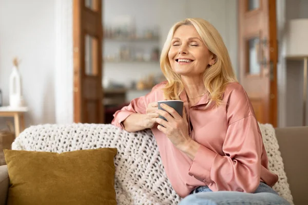 Portrait of joyful blonde senior woman holding cup of hot drink and enjoying her morning coffee, domestic comfort in spare time, posing in modern cozy living room indoors