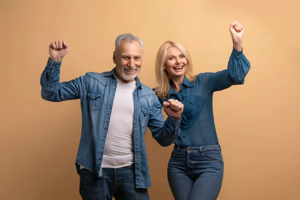 Thrilled happy retired couple elderly man and woman wearing casual outfit celebrating success together on beige studio background, clenching fists raising hands up and smiling