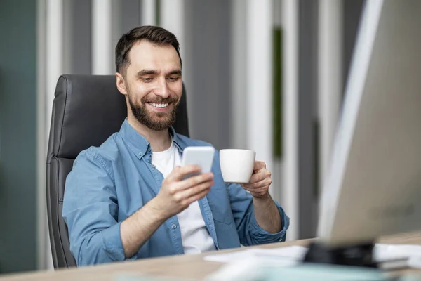 モダンオフィスでスマートフォンとコーヒーを飲む若いハンサムビジネスマン デスクで座っている笑顔の男性起業家 携帯電話でインターネットを閲覧し ホットドリンクを楽しむ — ストック写真