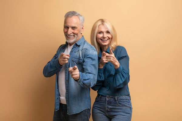 stock image Confident cool attractive caucasian elderly man and woman wearing casual denim outfit gesturing pointing at camera and smiling, isolated on beige background. Successful retirees