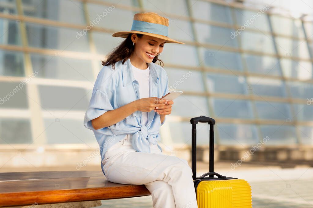 Smiling young european woman in hat with suitcase, typing on smartphone, enjoy trip at station, outdoor. App for tourist, travel with ticket, active lifestyle in summer with device