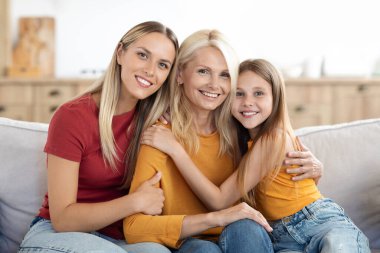 Neşeli küçük kız ve güzel milenyum kadını, mutlu anne, kız ve büyükanneyi kucaklayan evde koltukta oturan, kameraya sarılan ve gülümseyen, aile fotoğrafı için poz veren