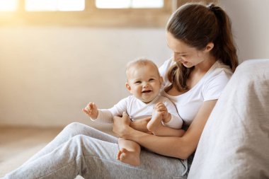 Genç, beyaz bir anne kucağında bebek, kucağında sevimli küçük bir kızı taşıyor, evde birlikte poz veriyorlar, güneş parlıyor. Çocuk bakımı, annelik ve annelik izni kavramı. Metin için boş boşluk
