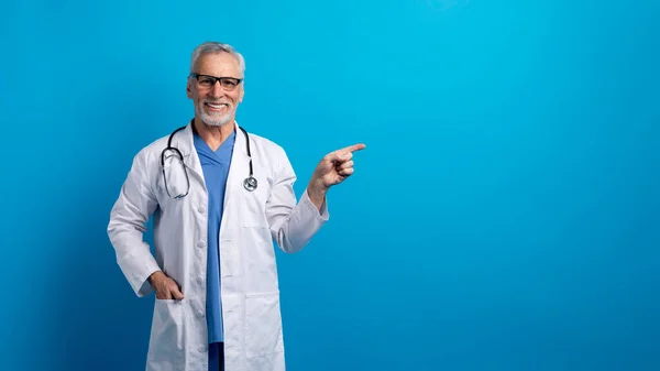 Healthcare, medical insurance. Cheerful handsome grey-haired bearded senior doctor in medical uniform and eyeglasses pointing at copy space for advertisement, isolated on blue background, panorama
