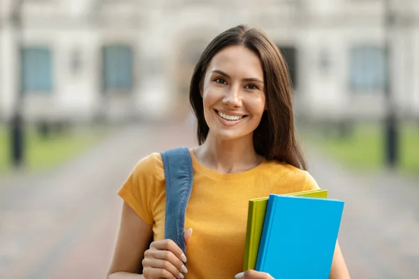 快乐的年轻女学生手握工作手册 背着背包来到学校大楼门口 美丽的千禧年淑女对着相机微笑 享受学习和教育计划 — 图库照片