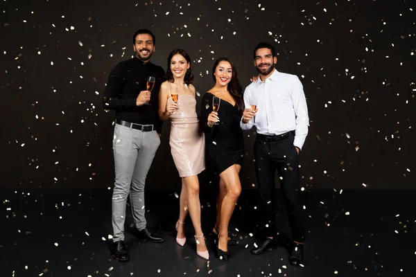 Happy beautiful international millennial friends couples in nice outfits have New Year party together, drinking champagne, enjoying falling confetti, black studio background. Christmas celebration