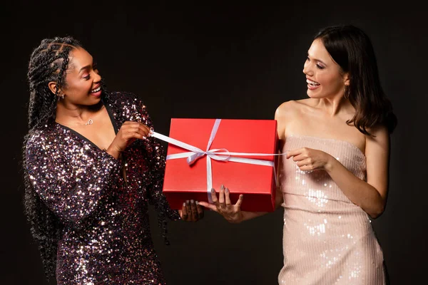 Joyful excited diverse multiethnic young ladies in nice outfits holding red gift box, opening Christmas present on black background. Happy cheerful girlfriends exchanging New Year gifts