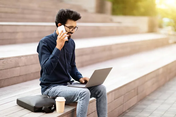 Telefonla bir fincan kahve ve laptop ile gülümseyen genç Hintli adam menejeri, şehirdeki merdivenlerde oturuyor, kopyalama alanı, güneş ışığı. İş, uzaktan iş, toplantı ve sağlık hizmetleri dışarıda.